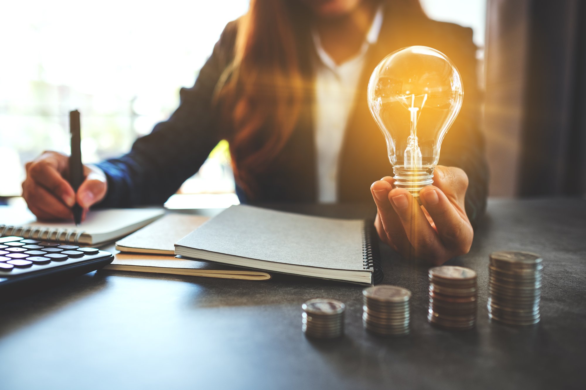 businesswoman-holding-a-lightbulb-while-taking-notes-stacked-coins